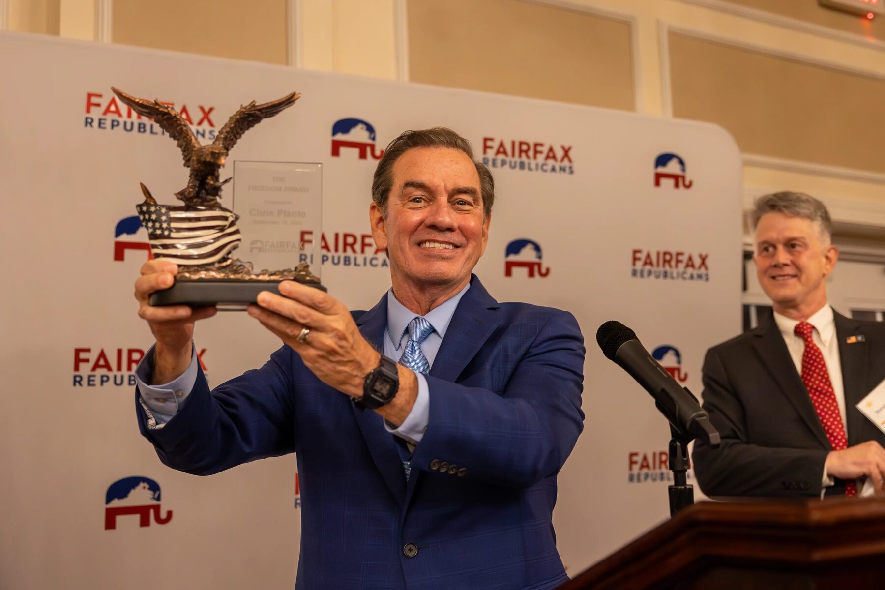 Chris Plante of News Talk 105.9 WMAL holds the Freedom Award — a bronze eagle over an American flag — aloft and smiles, in front of a Fairfax Republicans backdrop.