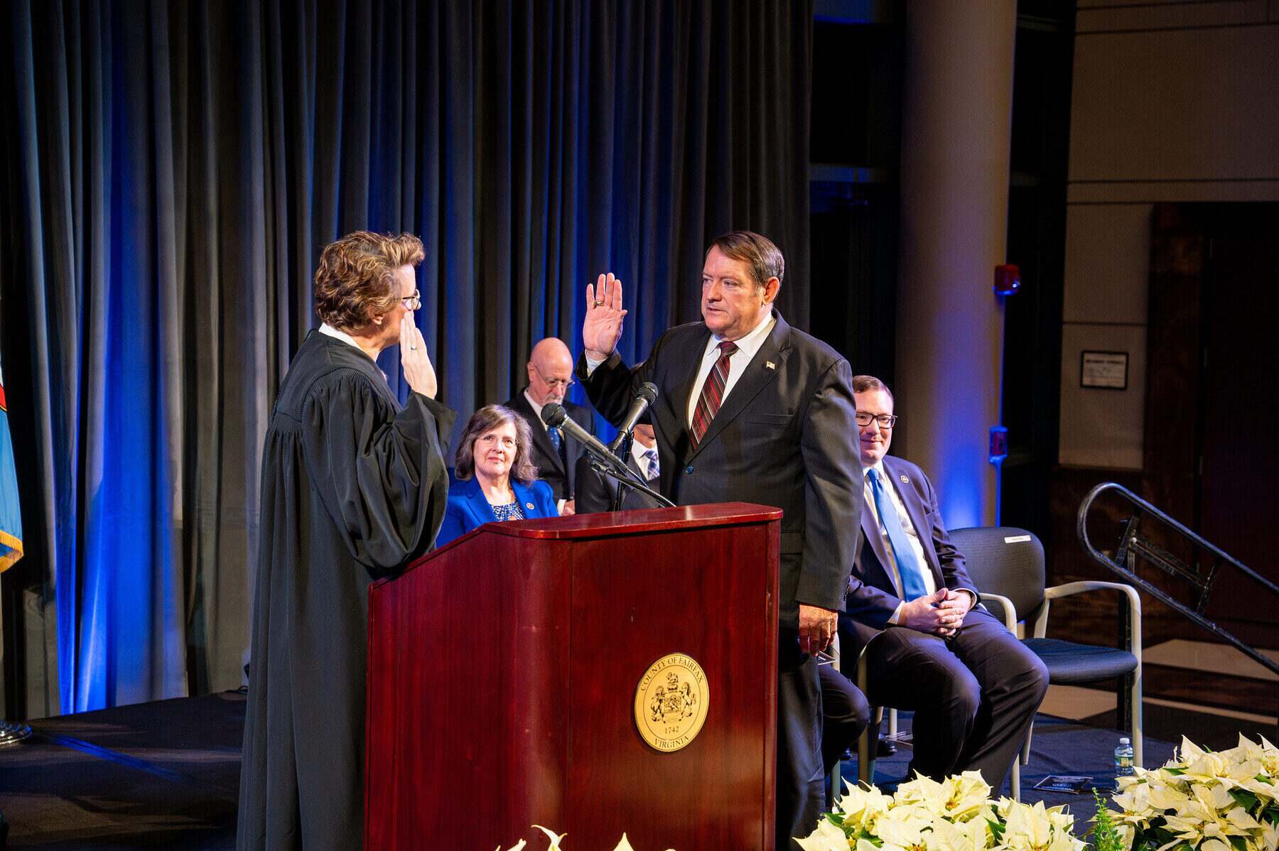 Supervisor Pat Herrity, right hand raised, takes the oath of office before a robed judge at a podium bearing the gold County of Fairfax seal, under blue civic lighting.