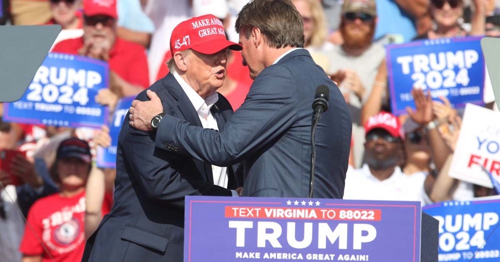 President Donald Trump, wearing a red Make America Great Again cap, embraces Glenn Youngkin on stage at a 2024 rally with Trump 2024 signs in the crowd.